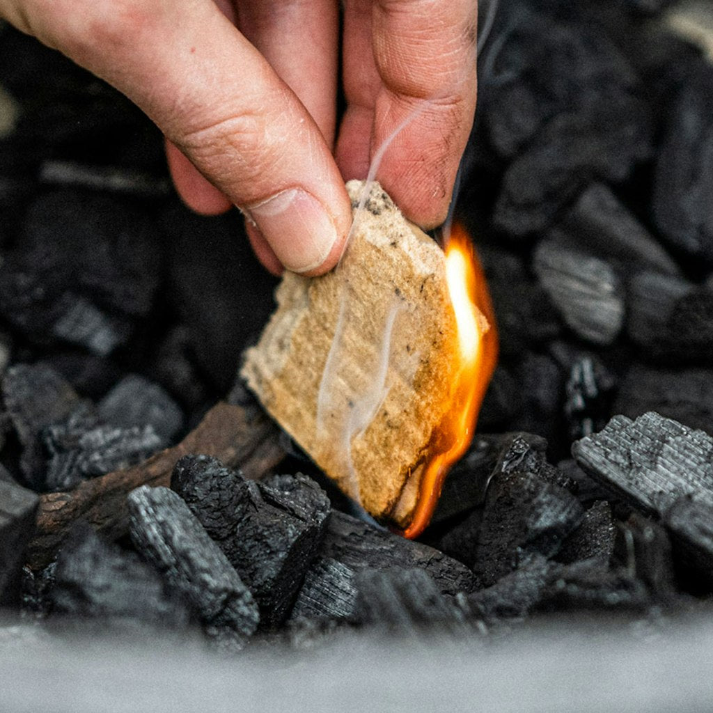 A close-up image of a person's hand holding a fire starter in a bed of charcoal with a small flame appearing from the charcoal.