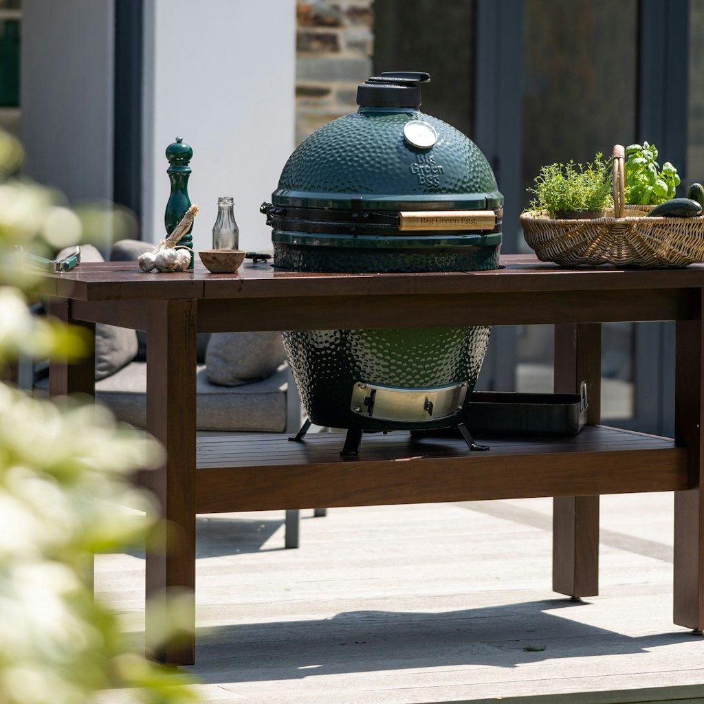 A Big Green Egg ceramic grill on a Premium Royal Mahogany Table with outdoor setting in the background.