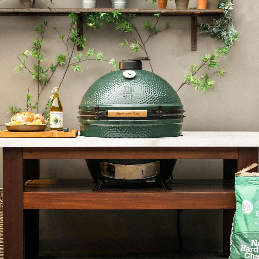 XL Big Green Egg grill on a wooden table with a bottle and a dish in the background, placed under a shelf with a plant.