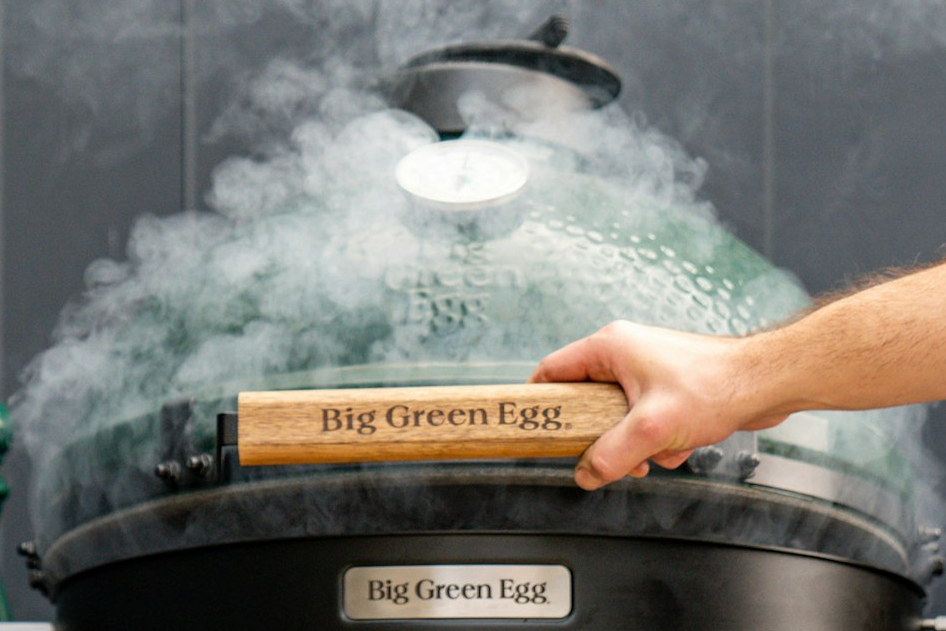 Person opening a Big Green Egg grill with smoke rising, brand name visible