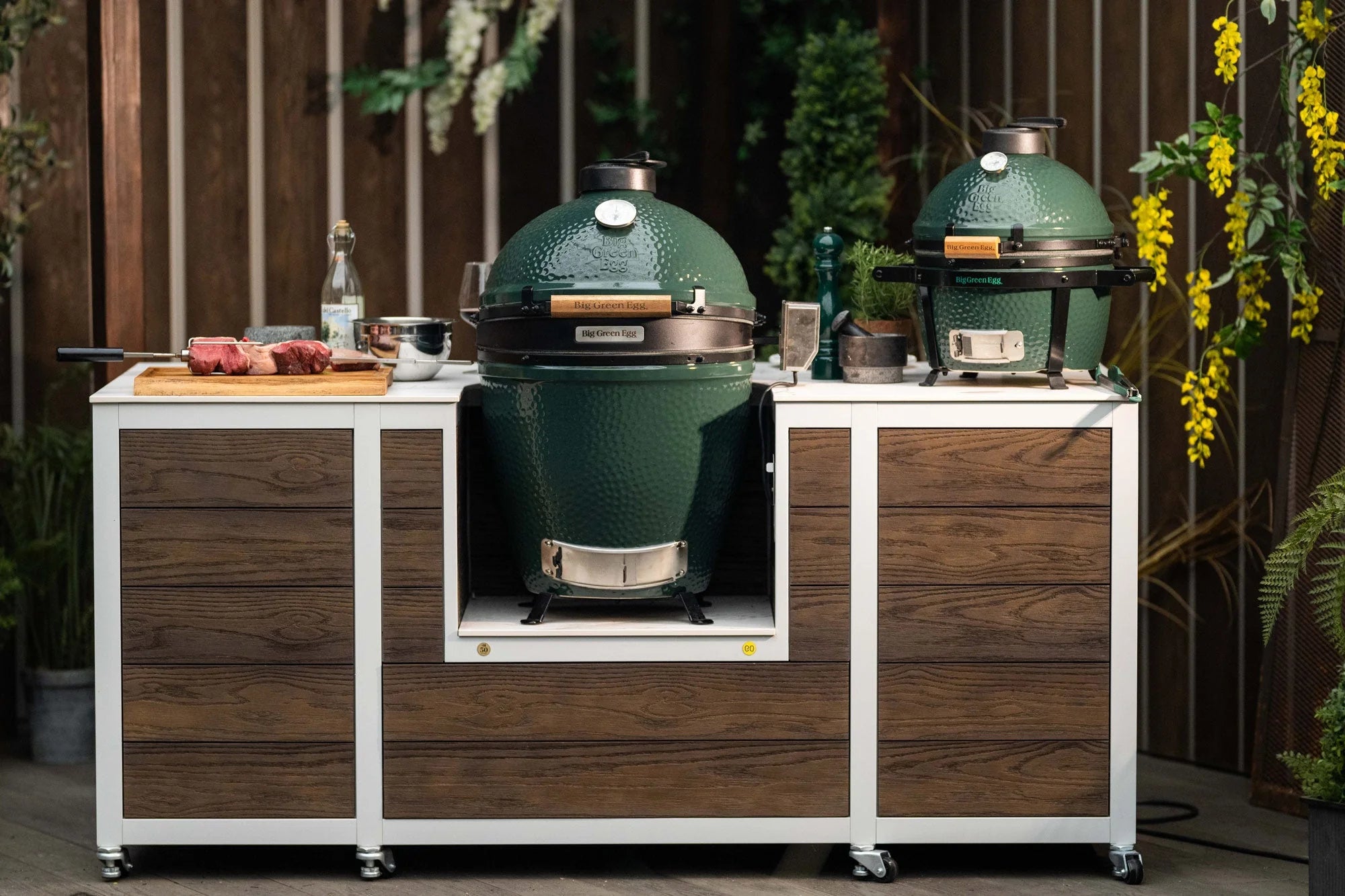 Two large green barbecue grills on a wooden and white cart outdoors.