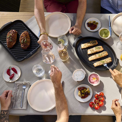 Overhead dining table scene with multiple people reaching toward food and drinks. On the table are two black oval griddles, grilled fish and steak, small plates of dips and condiments, glasses, plates, and utensils in a real indoor environment.