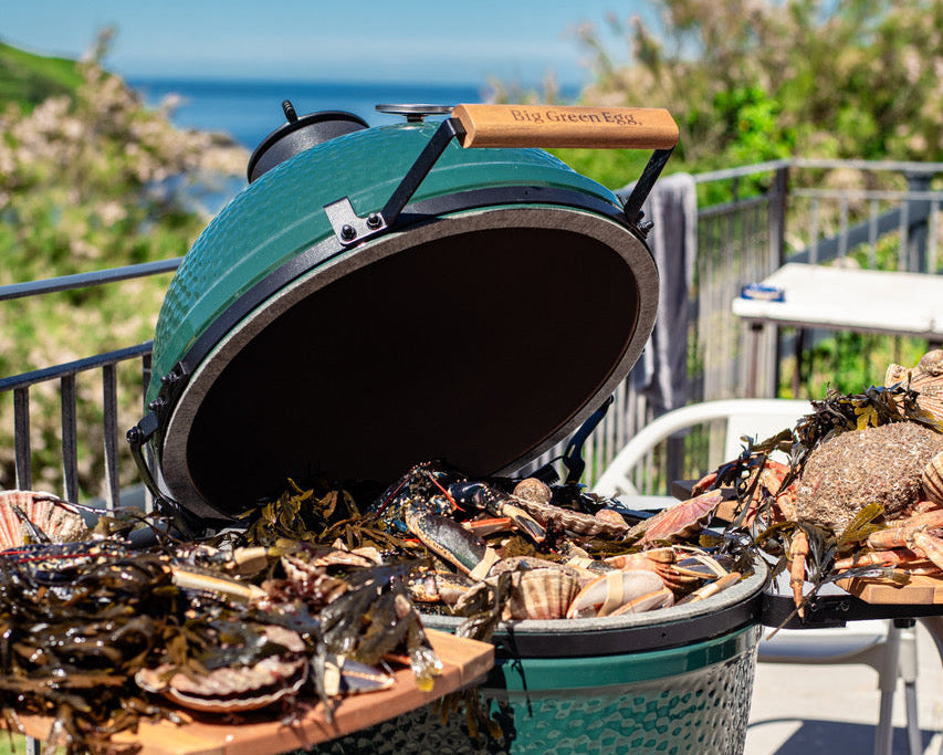 A green ceramic kamado grill open on an outdoor patio, filled with seafood including clams and mussels, with a scenic ocean and green hillside background.