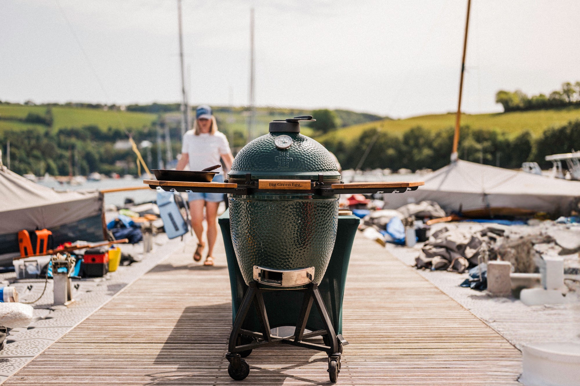 Large green barbecue grill on a wooden dock with people and boats in the background
