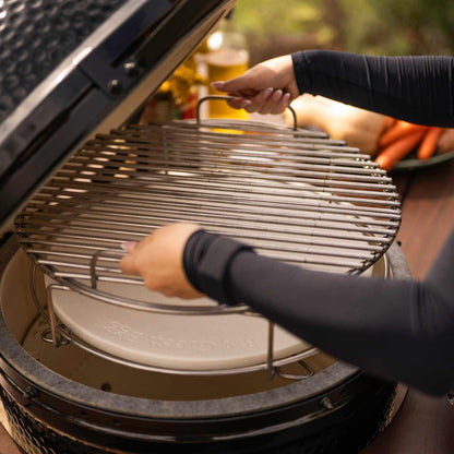 A person's hands adjusting a stainless steel grill grate inside a black ceramic kamado grill, with a white ceramic insert below and a blurred outdoor background. The Big Green Egg Dual Zone Kit.