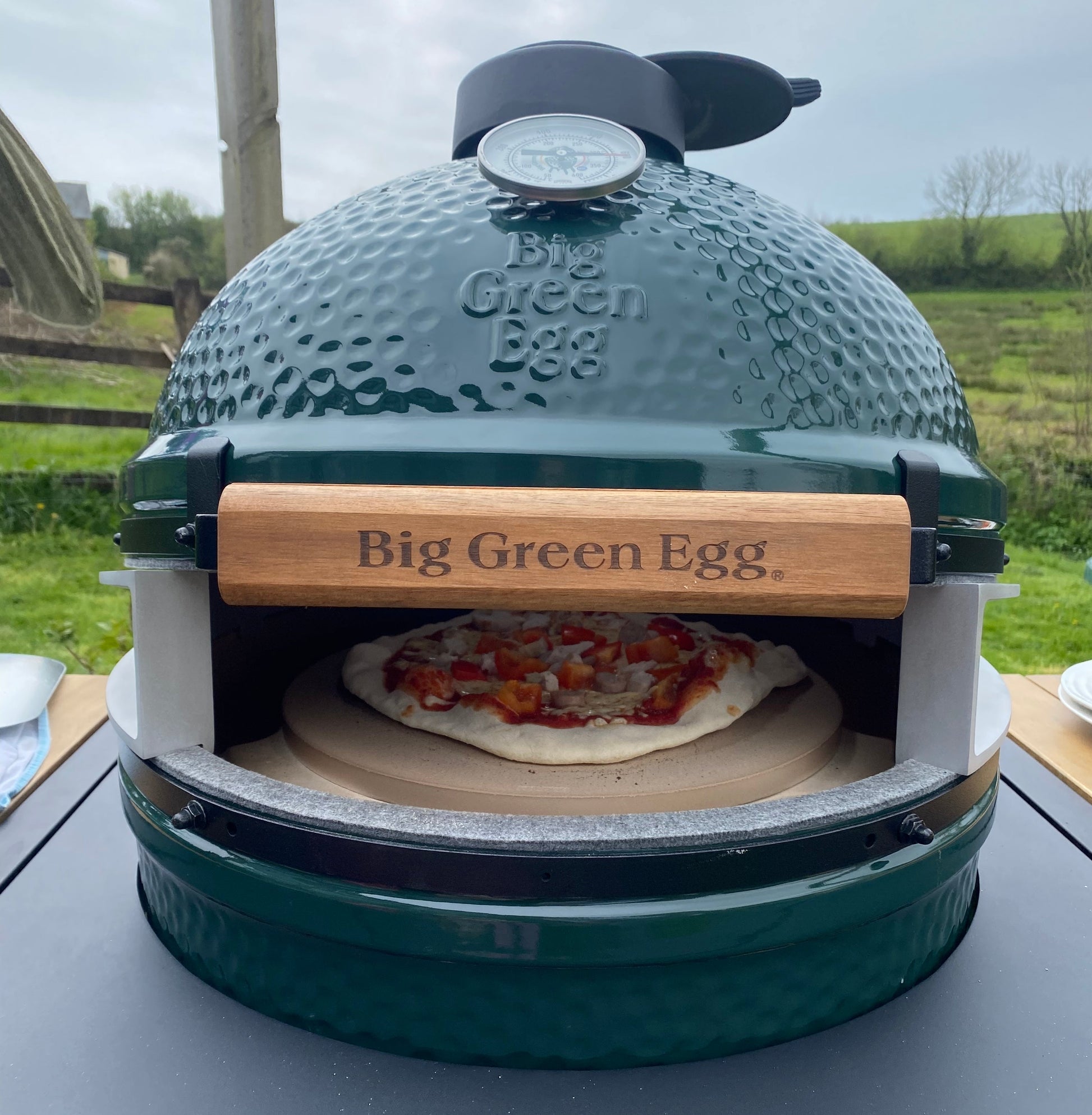 Big Green Egg outdoor pizza oven with a pizza inside on a table outdoors.