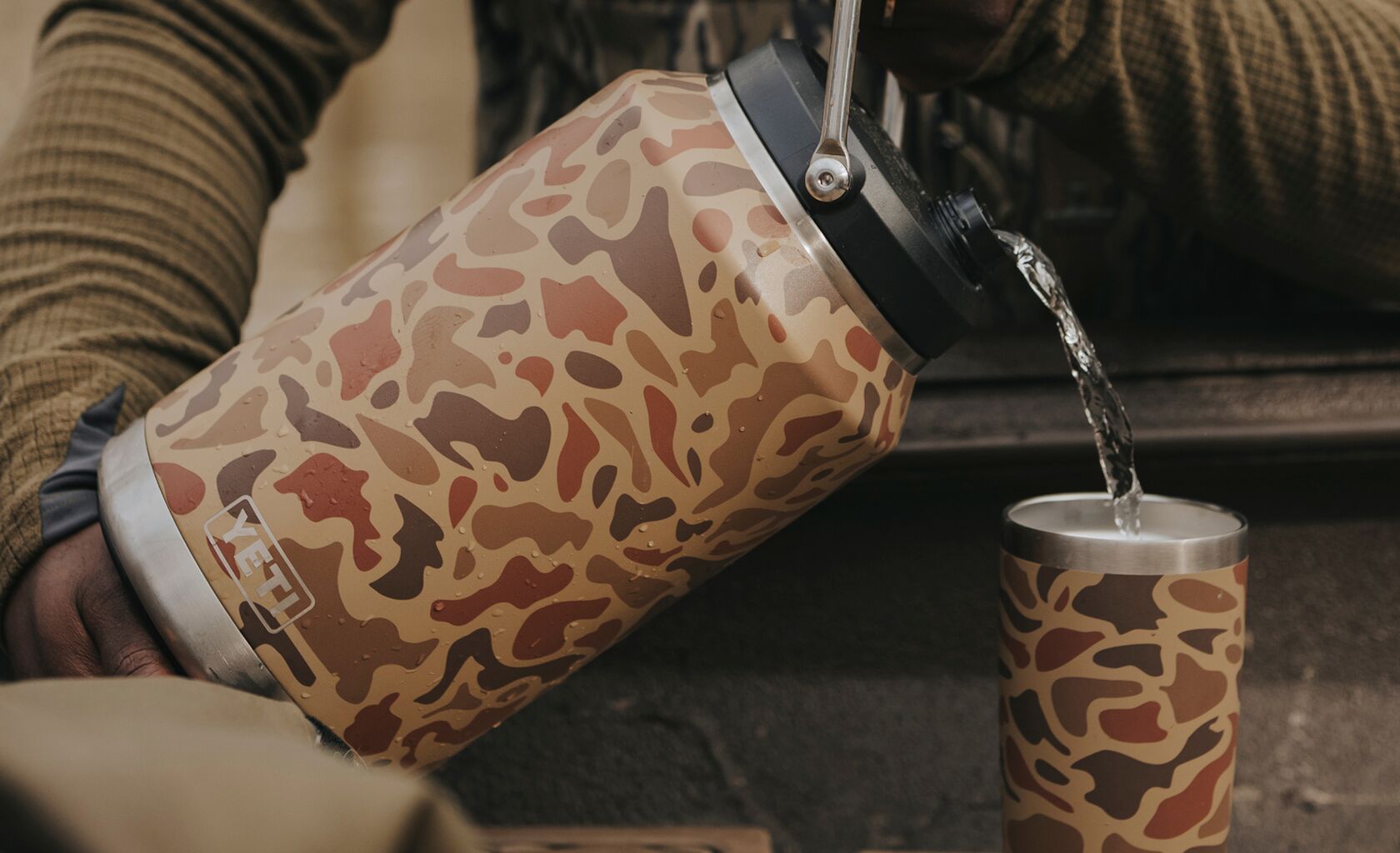 Camouflage-patterned tumbler being filled with water from another similar tumbler.