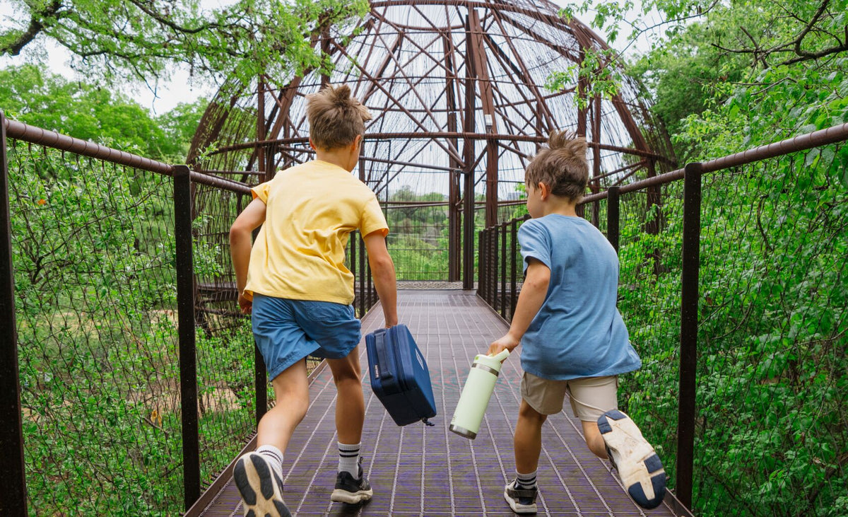 Two children running away from the camera on a metal walkway surrounded by green trees, one wearing a yellow shirt and blue shorts carrying a blue bag, and the other wearing a blue shirt and beige shorts carrying a white bottle.
