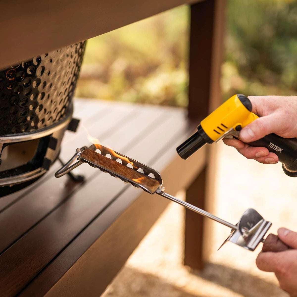 A close-up lifestyle shot of a person using a yellow TETRA portable torch lighter to ignite a piece of charcoal on a metal grill grate, set against a blurred outdoor patio background.