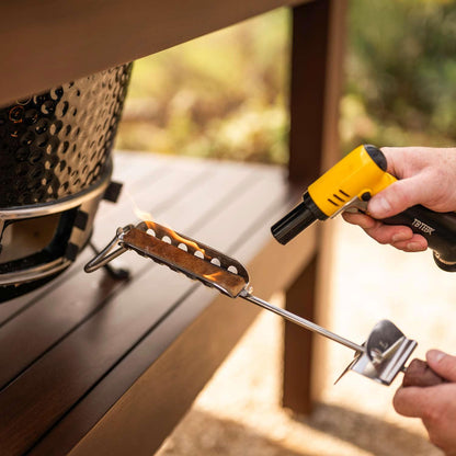 A close-up lifestyle shot of a person using a yellow TETRA portable torch lighter to ignite a piece of charcoal on a metal grill grate, set against a blurred outdoor patio background.