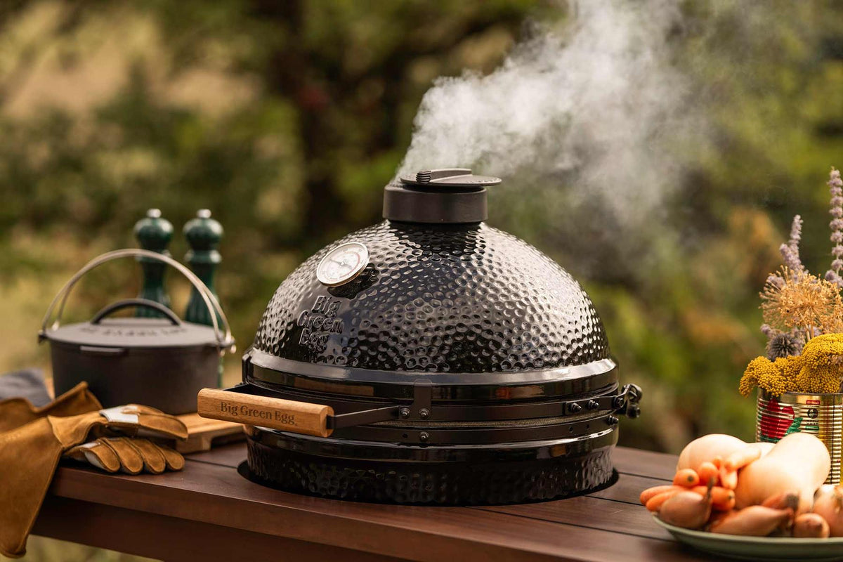 A black ceramic kamado grill with a wooden handle sits on a wooden table outdoors, with smoke rising from the top. A cast iron pot, leather gloves, and vegetables are arranged around it against a blurred green garden background.