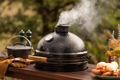 A black ceramic kamado grill with a wooden handle sits on a wooden table outdoors, with smoke rising from the top. A cast iron pot, leather gloves, and vegetables are arranged around it against a blurred green garden background.