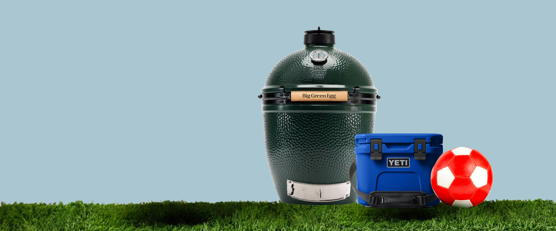 A green ceramic grill stands on grass next to a blue YETI cooler and a red-and-white soccer ball against a light blue background.
