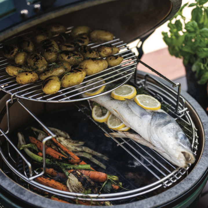 An open dark green ceramic kamado grill with multiple cooking racks. The top rack holds roasted baby potatoes, the middle rack holds lemon-sliced fish and vegetables, and the bottom rack holds carrots and greens over visible grill flames.