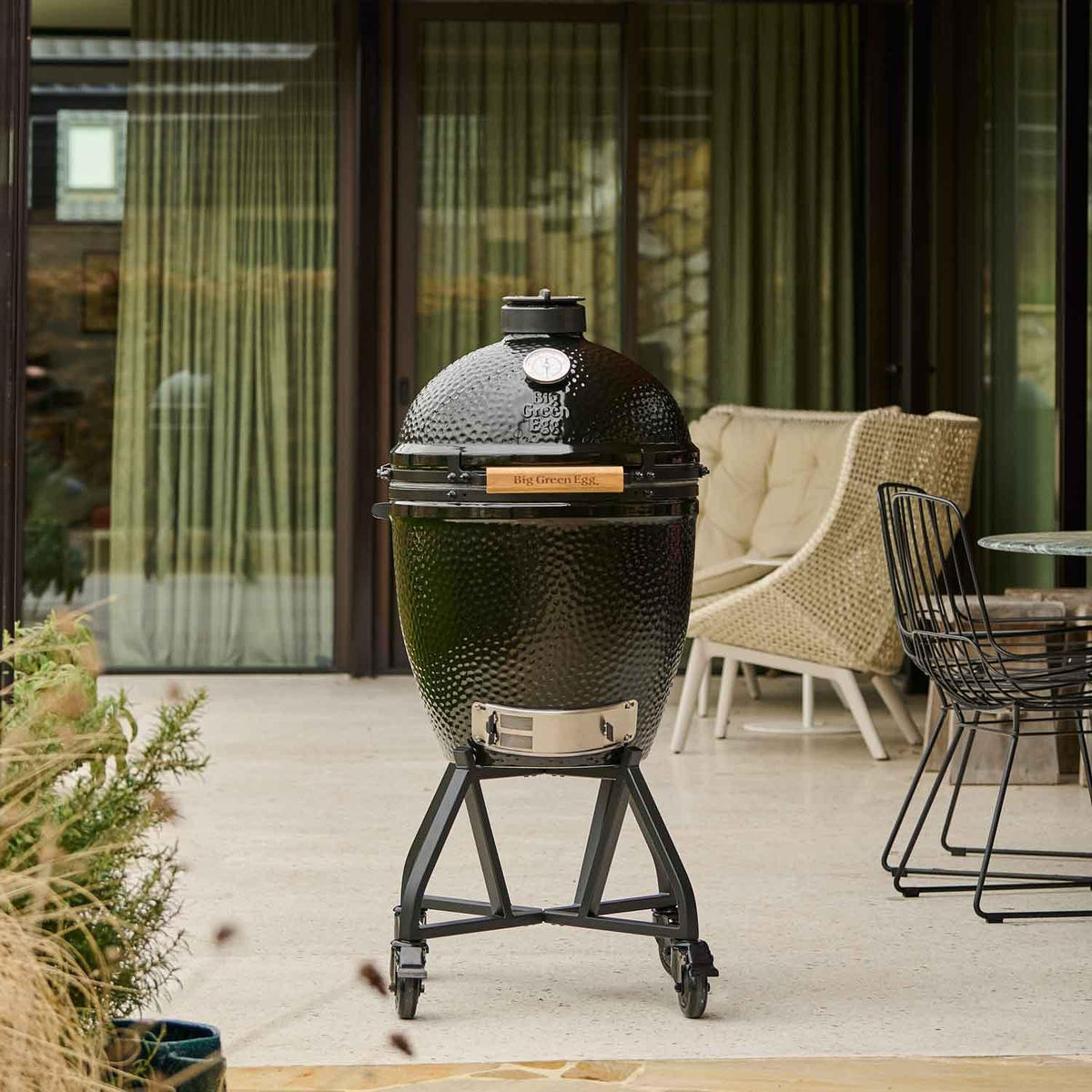 A black ceramic kamado grill on a matching wheeled stand, photographed on a patio in front of glass doors with chairs and curtains in the background.