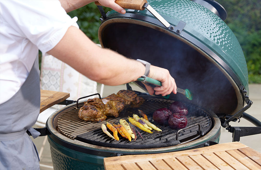 Person grilling vegetables and meat on a large green barbecue grill outdoors.