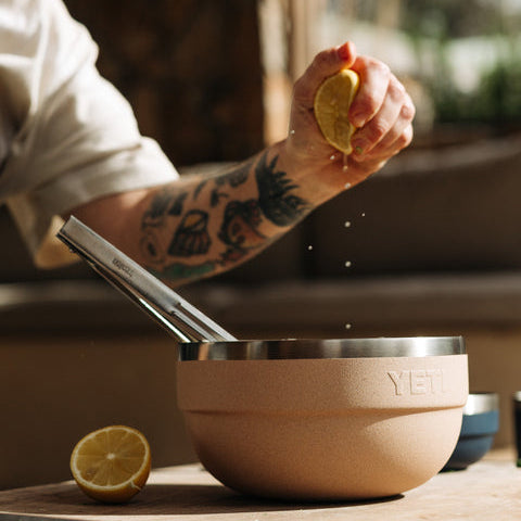 A person squeezes lemon into a YETI mixing bowl on a wooden table in a warm, rustic kitchen setting. The bowl has a matte tan exterior and a stainless steel rim, with the YETI logo embossed on the front.