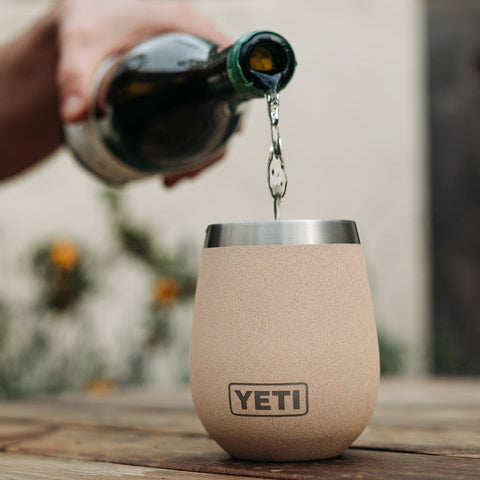 A YETI insulated stemless wine tumbler in a light beige textured finish sits on a wooden table, with a hand pouring a dark liquid from a bottle into the cup. The YETI logo is visible on the front of the tumbler, and a YETI logo watermark appears in the bottom right corner.