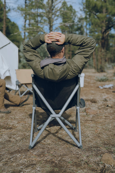 Man sitting in a camping chair with hands behind his head, surrounded by trees and a tent.