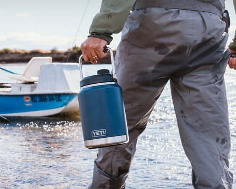 Person holding a YETI rambler one gallon jug by a body of water with text about the product's features.