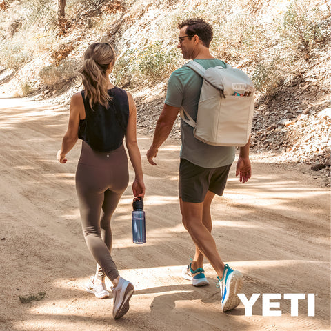 A man and a woman walking on a sunlit dirt trail wearing YETI backpacks, with the man's pack in light beige and the woman's pack in black. The man also wears a gray t-shirt and dark shorts, and the woman wears a black tank top and leggings.